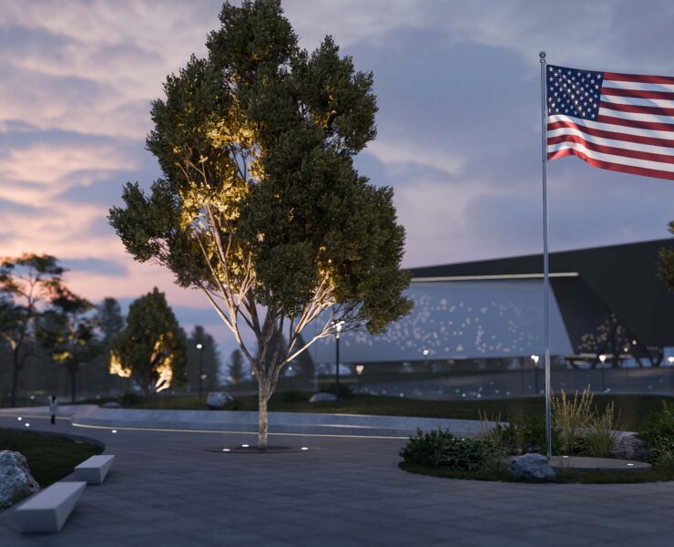 Alcon 9094 LED in-ground uplights illuminating an American flag on a flagpole in a landscaped plaza setting at dusk.