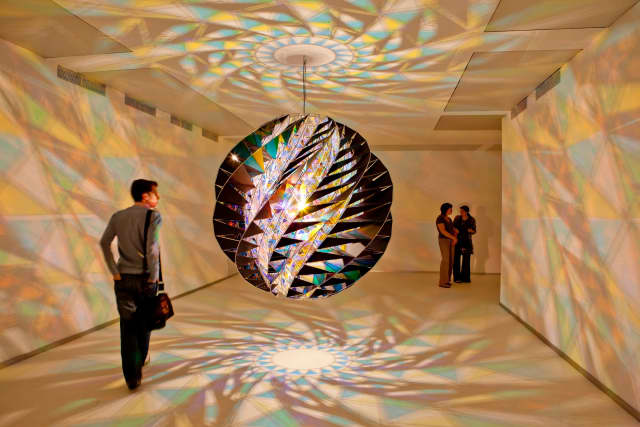Olafur Eliasson created the Lamp in Urban Movement in his exploration of spheres. A single light bulb hangs in a giant pendant light constructed of stainless steel, aluminum, black glass, and cyan and yellow filtered glass.
