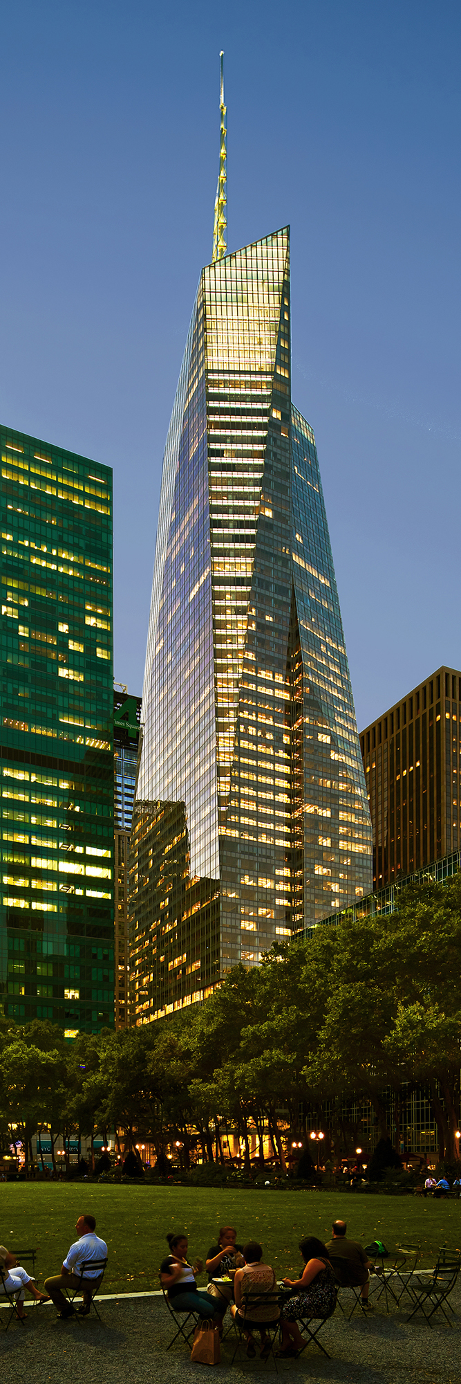 People sitting in Bryant Park at dusk with the illuminated Back of America Tower rising in the background.