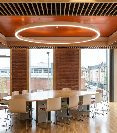 A ring pendant light suspends over a conference room table in a modern open floor plan office, anchored by adjustable aircraft cables.