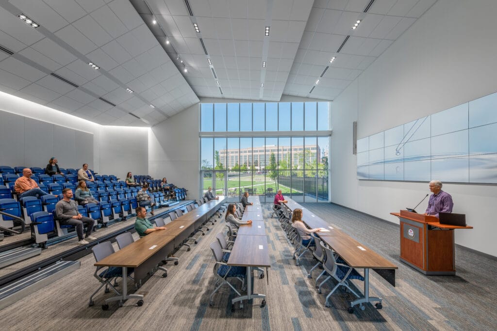 People attend a presentation in a lecture room with recessed lighting, indirect perimeter illumination and a large window wall.