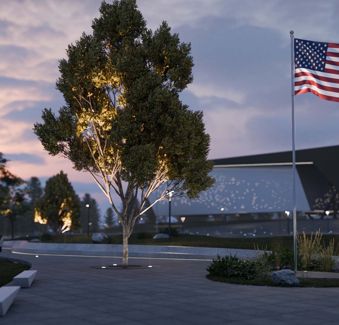 Alcon 9094 LED in-ground uplights illuminating an American flag on a flagpole in a landscaped plaza setting at dusk.