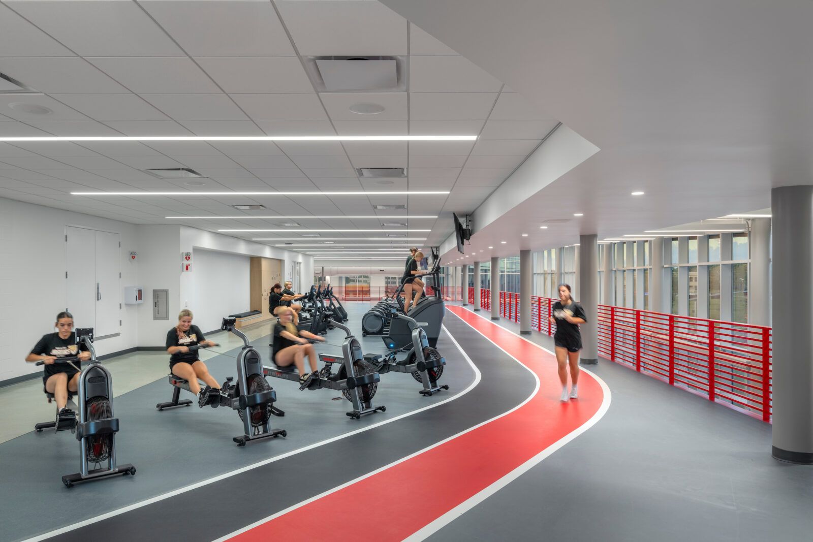 Students exercise along an indoor track illuminated by linear ceiling lights and daylight from perimeter windows.
