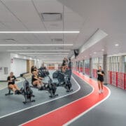 Students exercise along an indoor track illuminated by linear ceiling lights and daylight from perimeter windows.