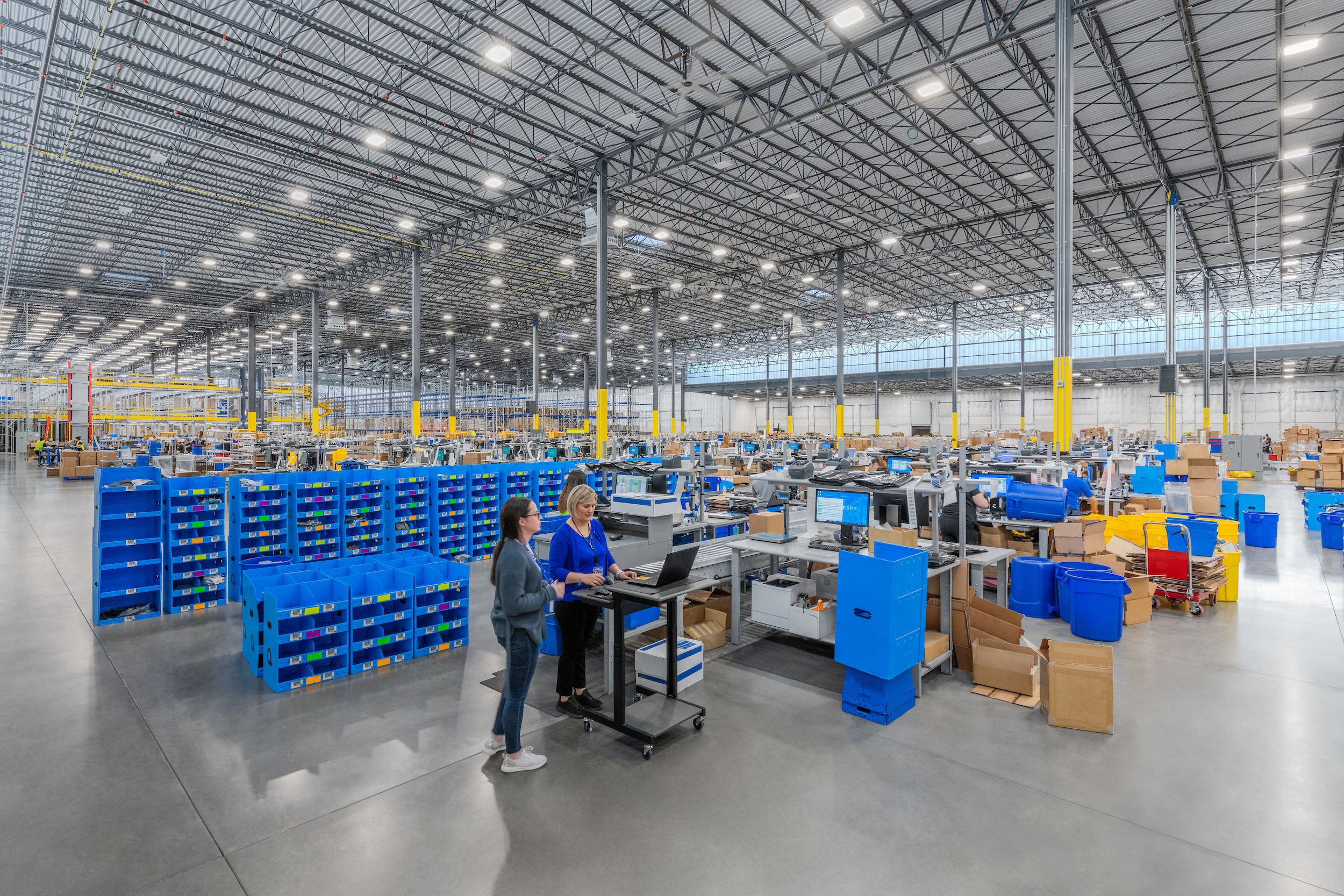 Workers in a large distribution center illuminated by LED high bay fixtures and daylight entering through clerestory windows.