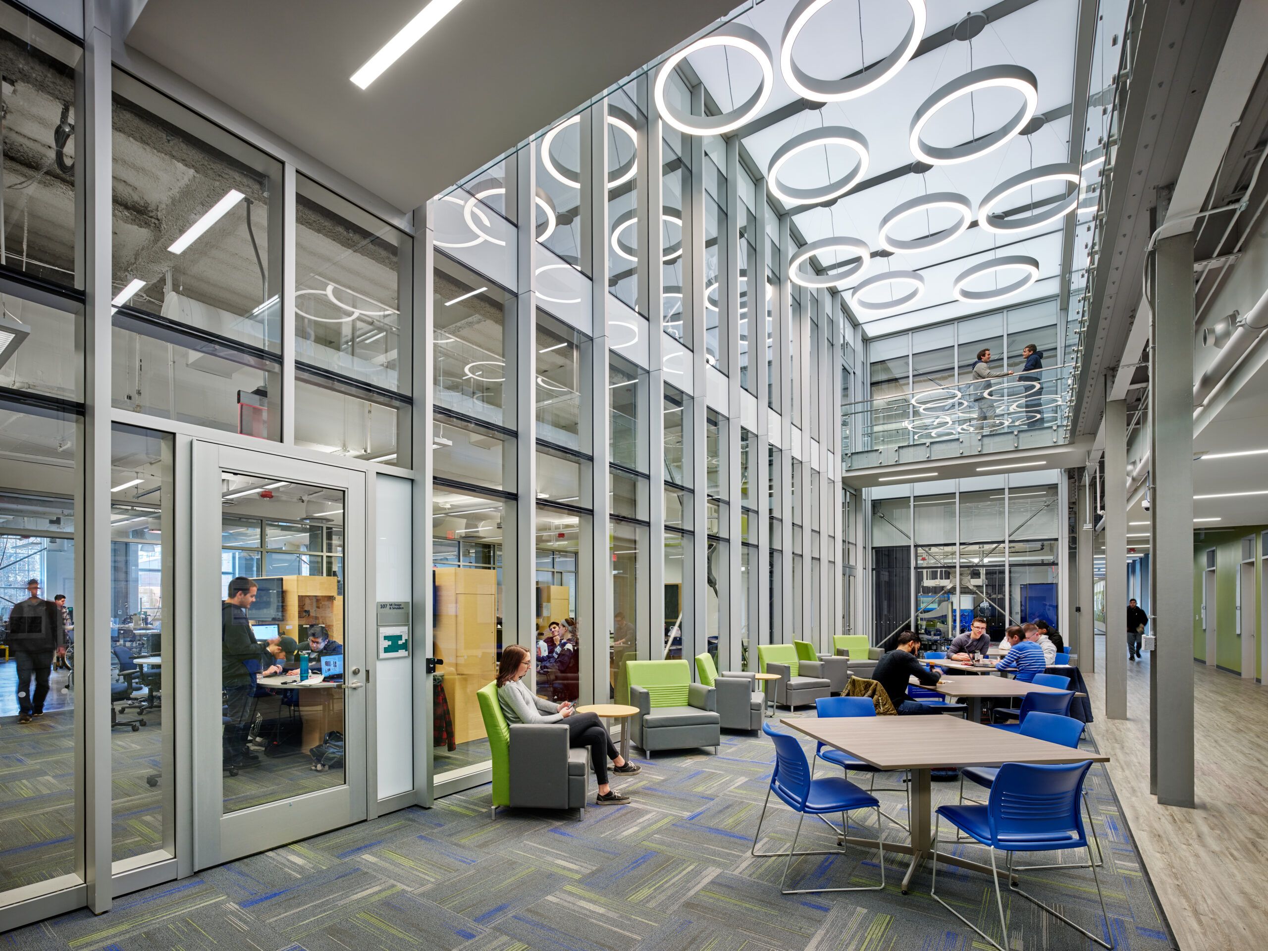 Students gather in a daylight-filled education atrium illuminated by suspended ring lights and linear architectural recessed lighting.