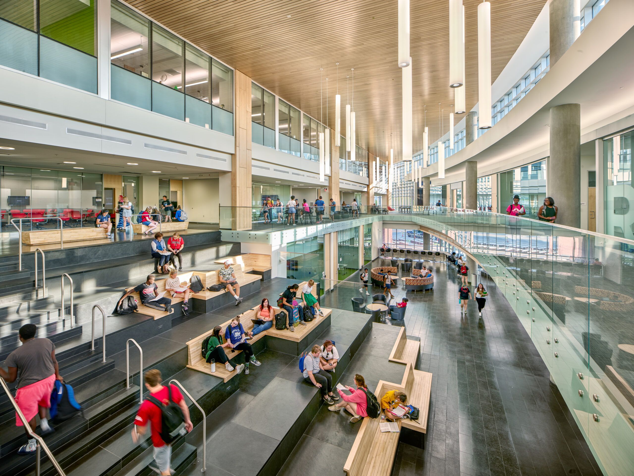 Students occupy a multilevel atrium with extensive glazing, a wood ceiling and suspended cylinder pendant lights.
