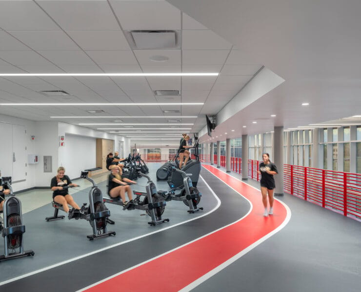 Students exercise along an indoor track illuminated by linear ceiling lights and daylight from perimeter windows.