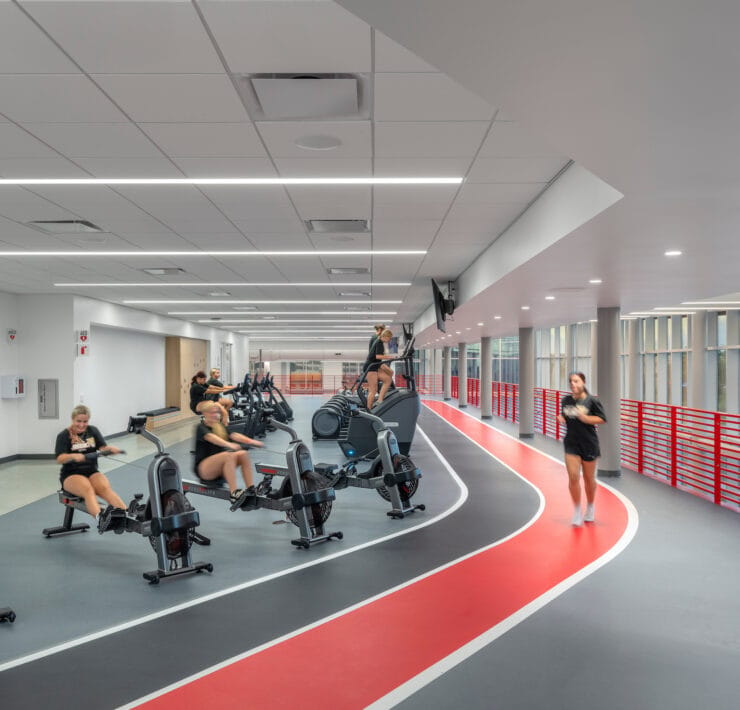 Students exercise along an indoor track illuminated by linear ceiling lights and daylight from perimeter windows.