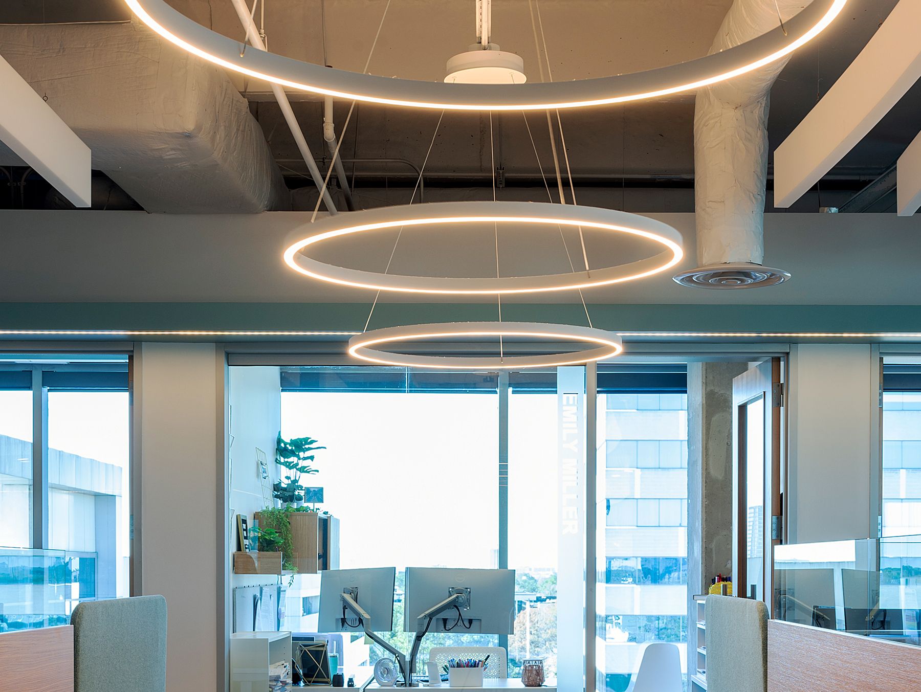 Three round LED pendant lights with a white finish illuminate the main walkway of an open floorplan office area, suspended by adjustable aircraft cables.