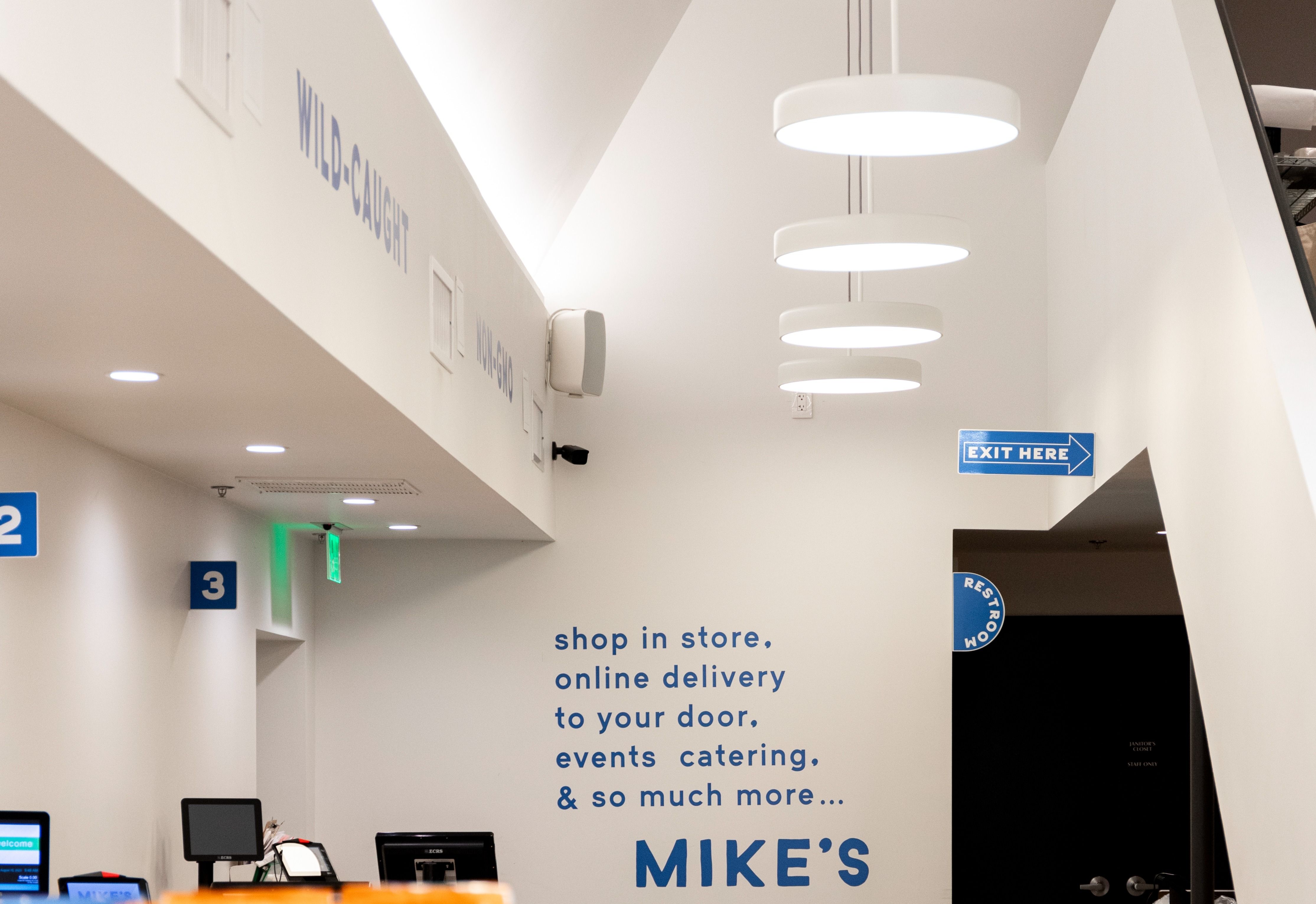 White round panel pendant lights suspended from a sloped ceiling on a white stem mounting, providing the customer clarity in wayfinding from the grocery store entrance. 