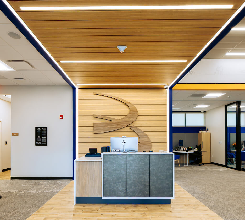 Linear recessed lighting mounted in a wood ceiling lights the way to the welcome desk of a Colorado credit union.