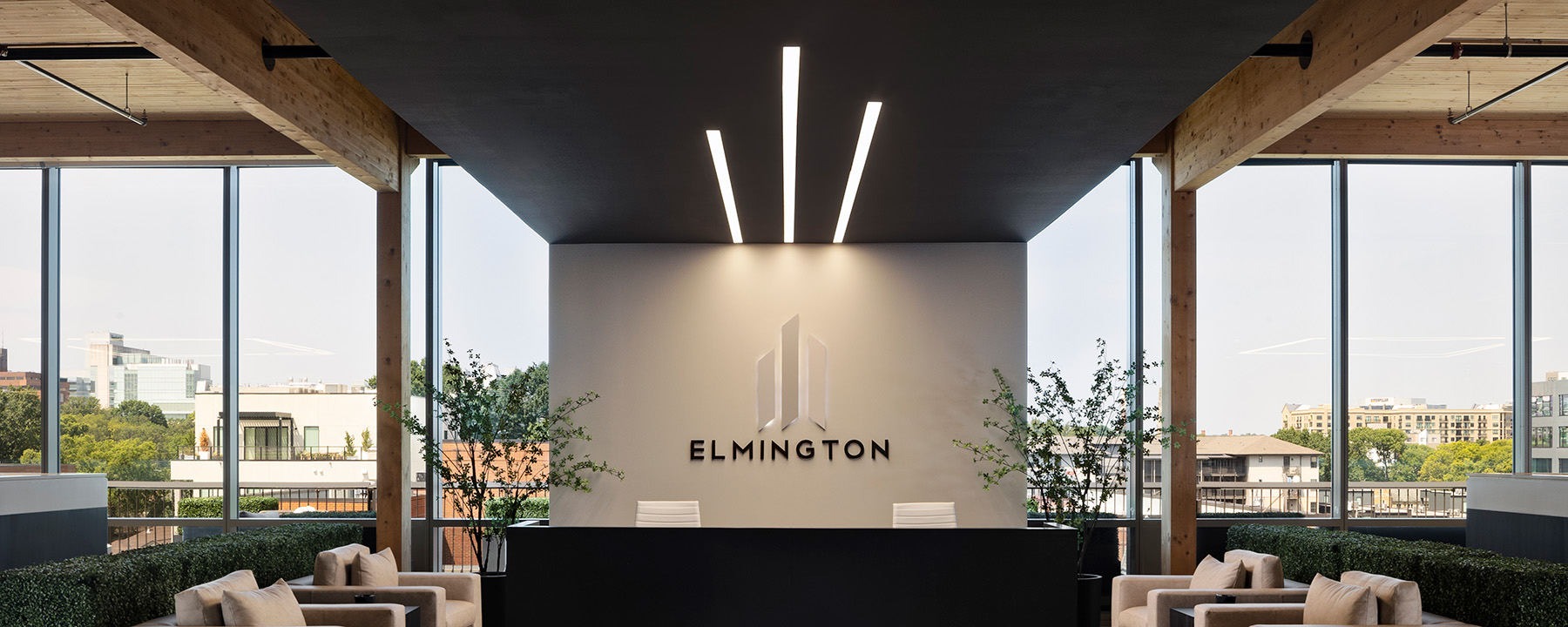 Three linear ceiling lights of different lengths align with and match the three towers featured in the Elmington logo behind the office lobby desk, creating a lighting design detail that reinforces branding.
 