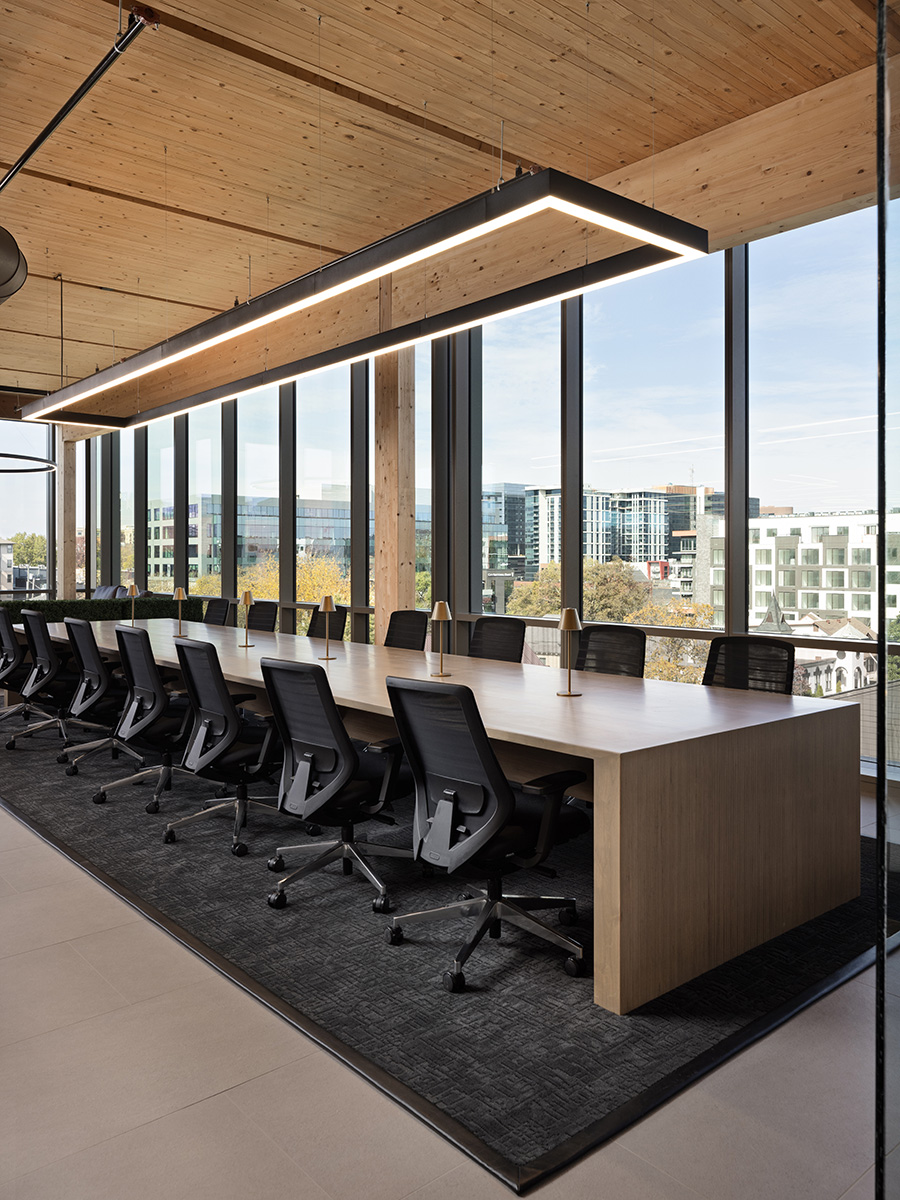 A rectangular LED pendant downlight suspended over a long conference room table in a corner office conference room with ample lighting through floor-to-ceiling windows.
 