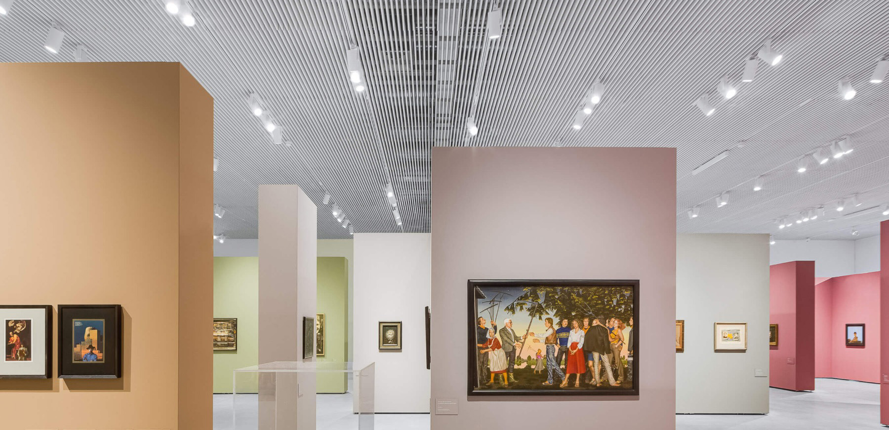 White cylinder LED track lights illuminate wall art in a gallery, mounted on a track channel recessed in a slatted wood ceiling.