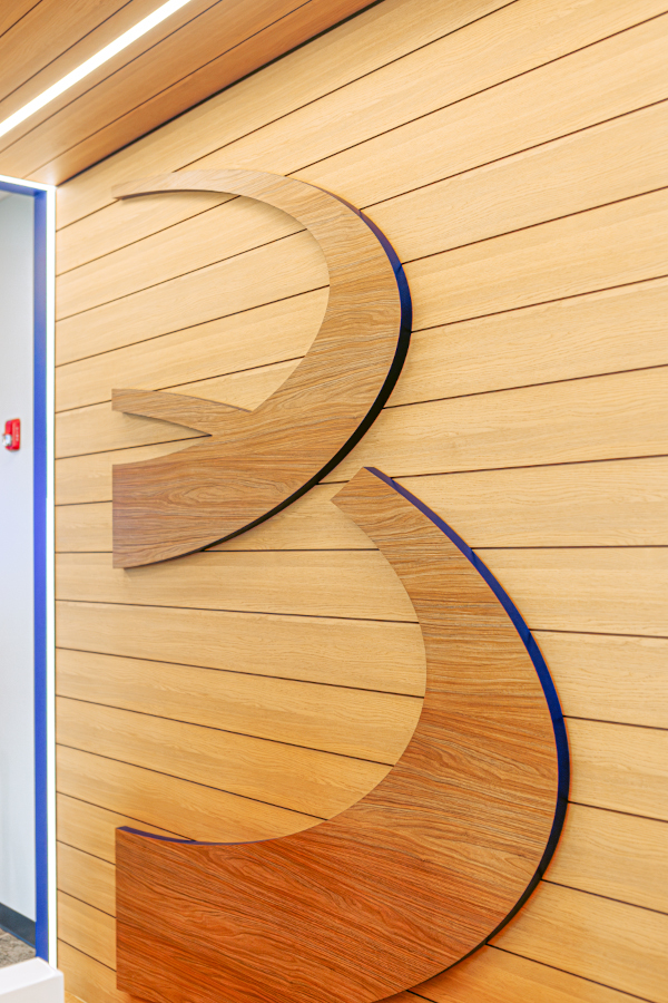 An integrated T-bar linear light fit in a slatted wood ceiling illuminates a wood panel Bellco logo wall detail.