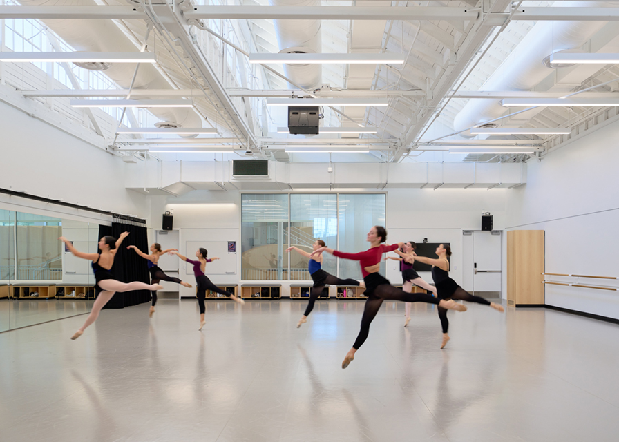 Slim linear pendant lights hang from a sloped ceiling over a dance studio classroom