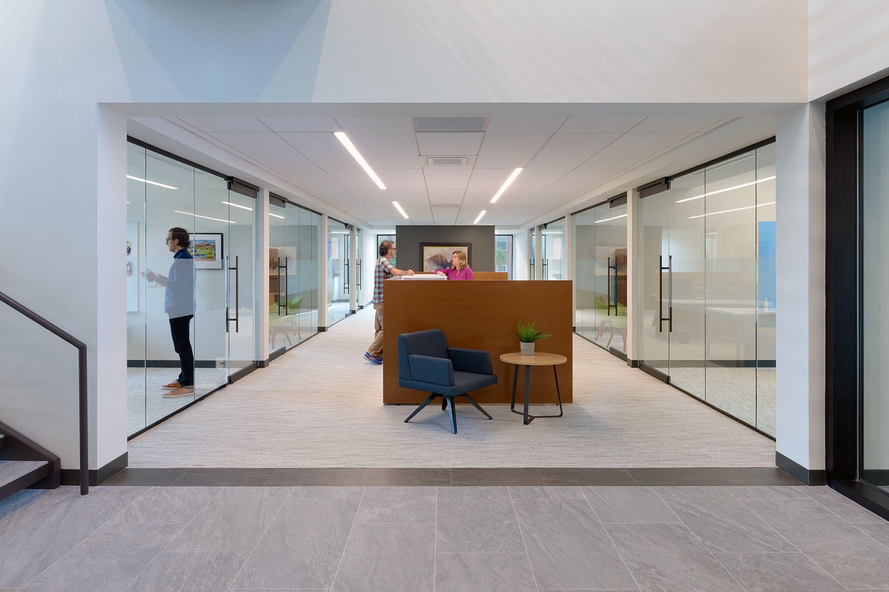 Linear recessed lights line a grid ceiling in a team collaboration area between offices with sliding glass fronts.