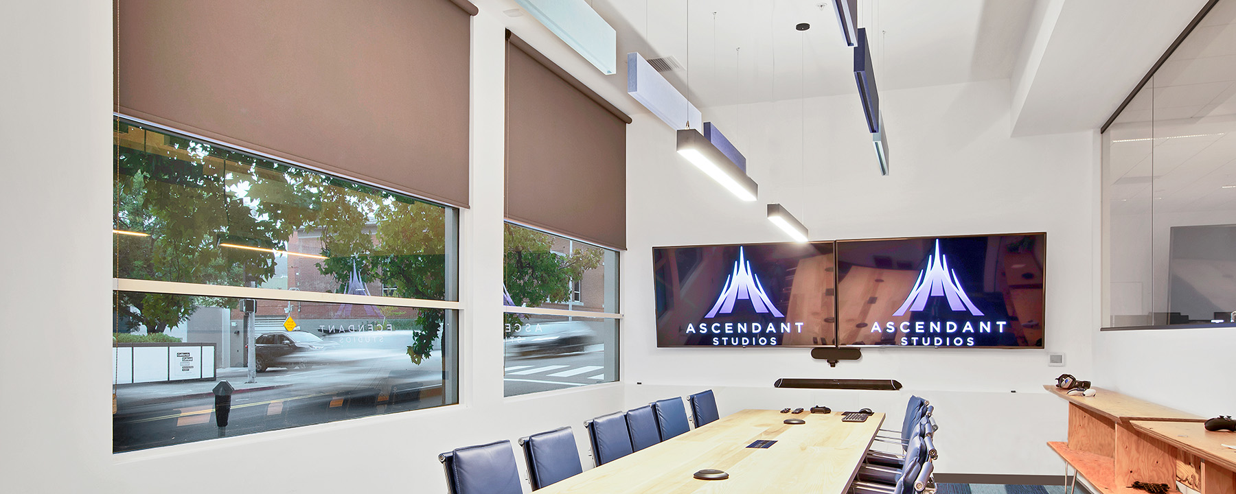 Linear suspension lighting hanging over a conference room table at Ascendent Studios balances with natural light coming through the windows. Shades that can be drawn for video viewing during meetings.